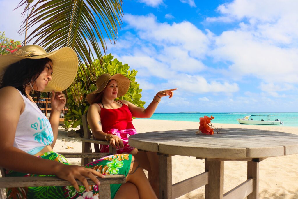 Two women sitting on wooden chairs with a table infront of them at the beach